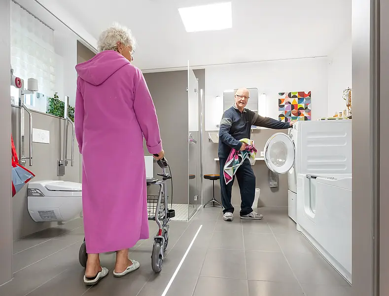 A woman with a walker wearing a bathrobe and a man operating a washing machine. 