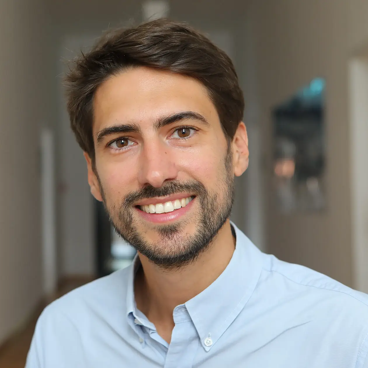 A portrait photo of Cosimo Posth, a white man with dark brown hair and a short beard, facing the camera widely smiling, wearing a light blue button down shirt.