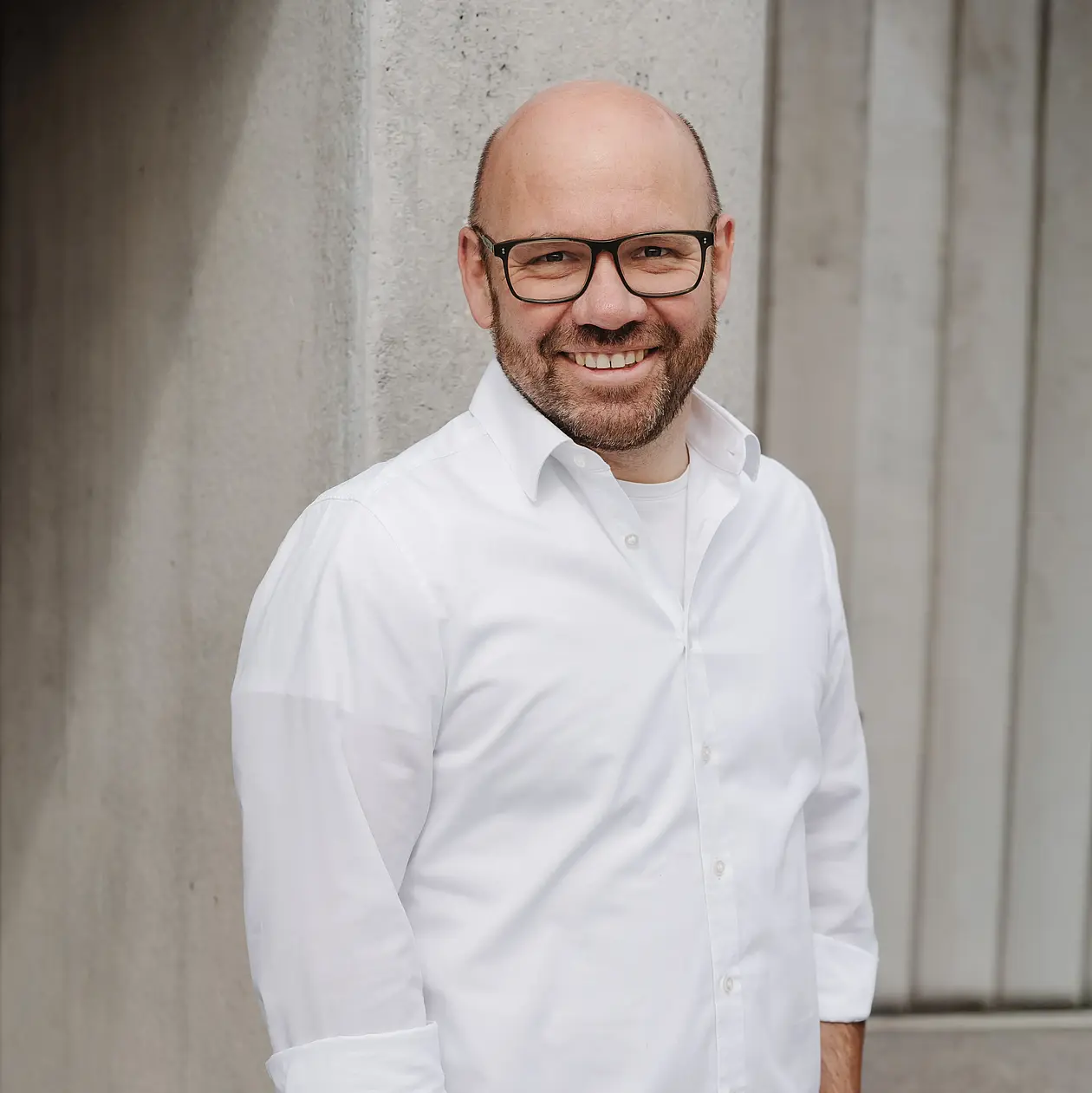 A photo of Sven Nansen, a white middle aged bald man wearing square glasses and smiling into the camera. He is wearing a white button down shirt and has a short beard stubble. He is standing in front of modern concrete architecture. 
