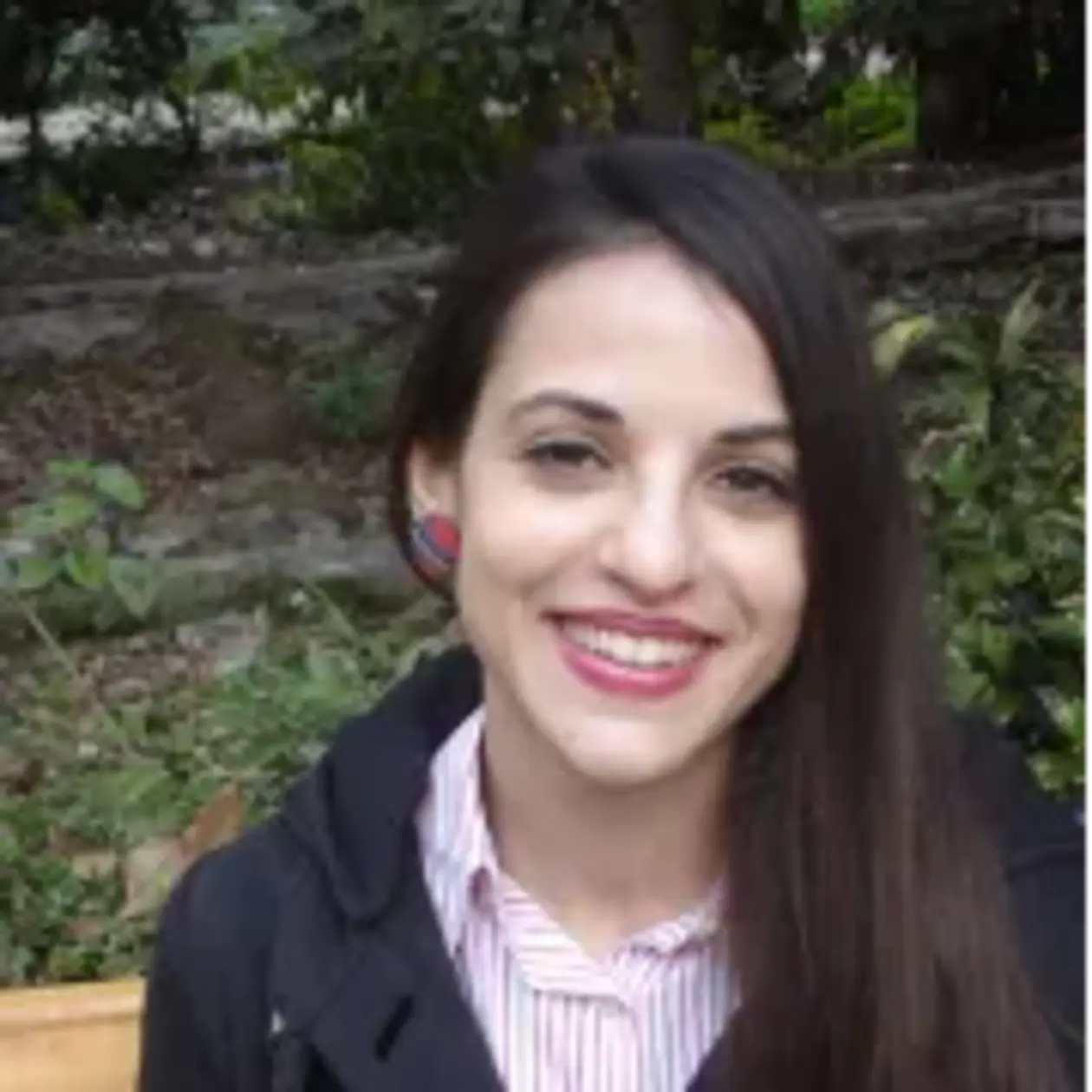 The photo shows Stella Kyrikou, a white woman with long straight black hair from the front smiling at the camera. She wears a dark blue cardigan, a blouse with thin white and red stripes and red earrings. 