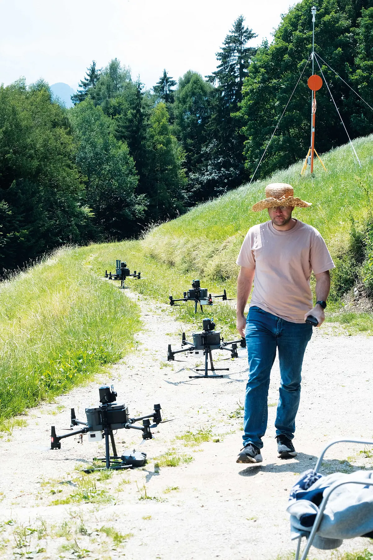 Ein Mitarbeiter läuft einen Weg hoch, rundherum Wiesen und auf dem Weg hinter ihm sind vier Drohnen zu sehen. 