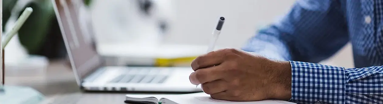 Auf diesem Symbolbild ist eine Person zu sehen, die vor einem Laptop sitzend, in ein Notizbuch schreibt. Dieses Bild steht für die E-Learning Angebote des Instituts.