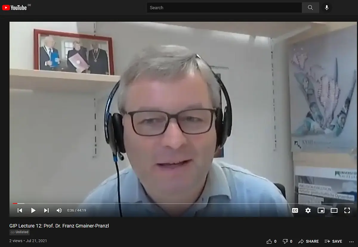 A man with gray hair and headset talking into a webcam