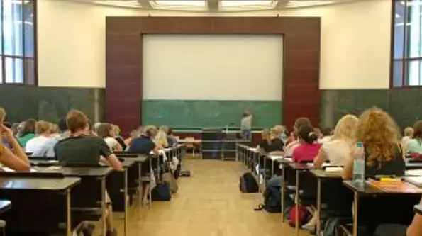 Links und rechts vom Gang sitzen Schüler*innen an Holztischen. Vorne schreibt ein*e Lehrer*in auf eine grüne Tafel.