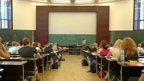 Links und rechts vom Gang sitzen Schüler*innen an Holztischen. Vorne schreibt ein*e Lehrer*in auf eine grüne Tafel.