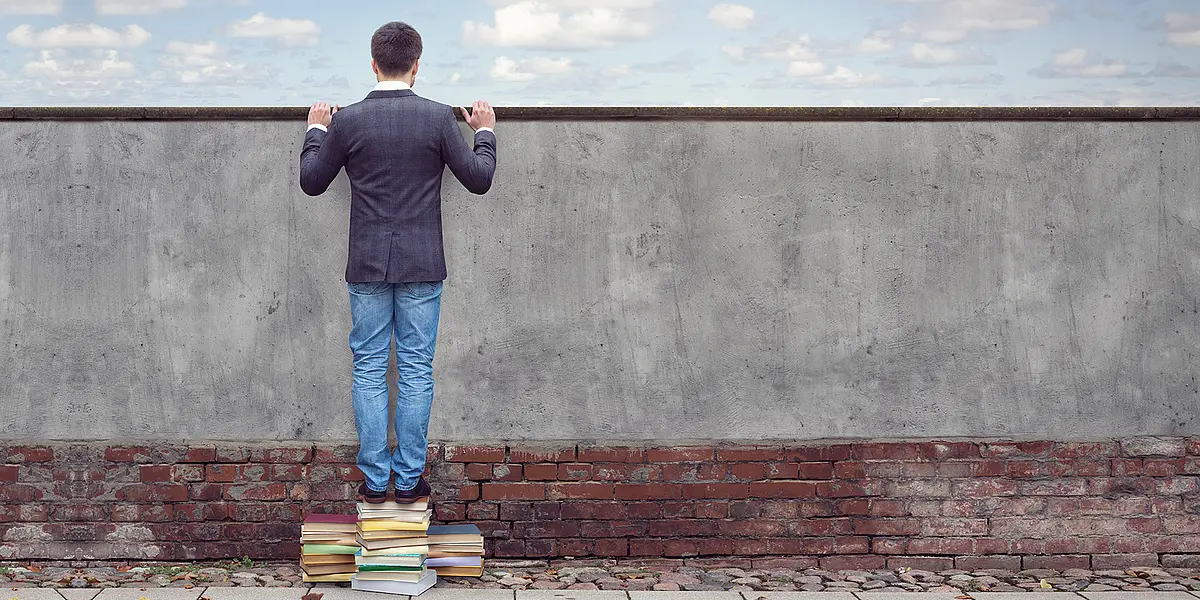 Ein Mann steht auf einem Stapel Bücher und kann dadurch über eine Mauer blicken. Was er sieht, wissen wir nicht. 