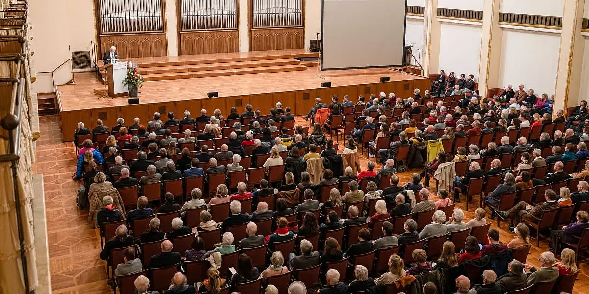 Der gute gefüllte Festsaal der Universität. Im Vordergrund die Sitzreihen mit dem Publikum mit dem Rücken zur Kamera. Im Hintergrund auf der Bühne am Redepult ist ein Redner erkennbar. Dahinter die großen Orgelpfeifen des Festsaals.