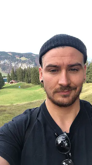 A Selfie by David Boysen, a white man with a short dark beard wearing a black beanie and a black shirt. He wears dark studded earrings and has black sun glasses hanging from the neckline of his shirt. He is standing in an alpine landscape with a high mountain and evergreen trees in the background. 