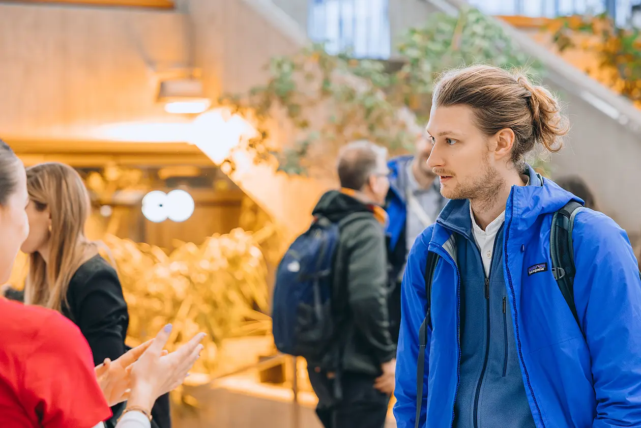 Ein Student mit blauer Jacke ist anderen Personen zugewandt. Er befindet sich bei einer Veranstaltung. 