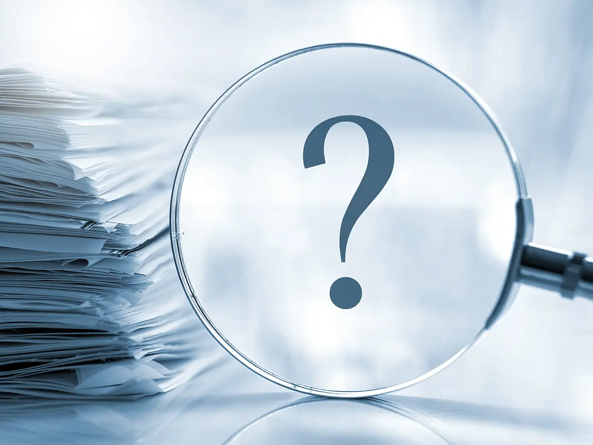 Picture with a stack of documents, in front of it a magnifying glass with an enlarged question mark
