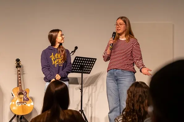 Two women standing on a stage while one of them is talking to a crowd of people using a microphone