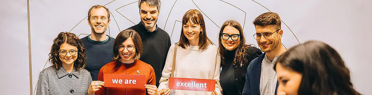 Ein Gruppe von 8 Personen schauen in die Kamera und halten Schidler hoch mit dem Text "We are excellent"