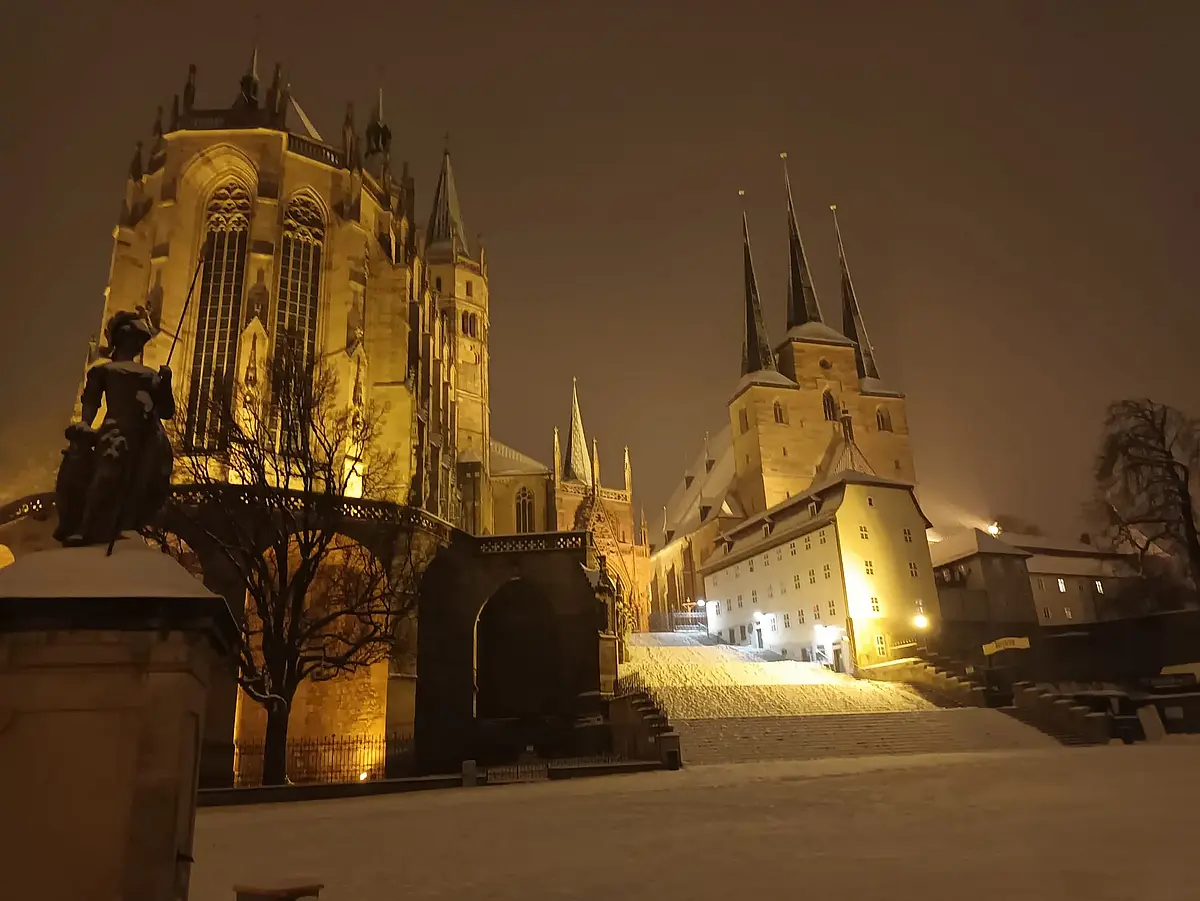 Auf dem Bild ist der Erfurter Dom bei Nacht in winterlicher Atmosphäre zu sehen. Die Straßenlaternen tauchen die Szenerie in ein warmes, orangefarbenes Licht