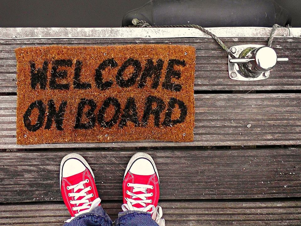 Zwei Füße auf Hafenplanken vor Fußmatte mit der Aufschrift "Welcome on Board"