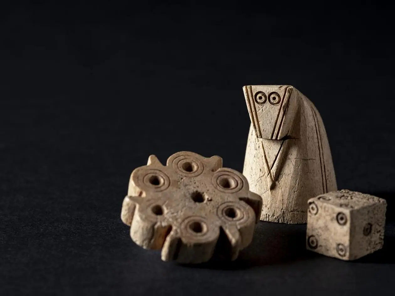 Das Foto zeigt die Schachfigur, den Spielstein und den Würfel vor schwarzem Hintergrund. Alle drei Funde sind beigefarben. 