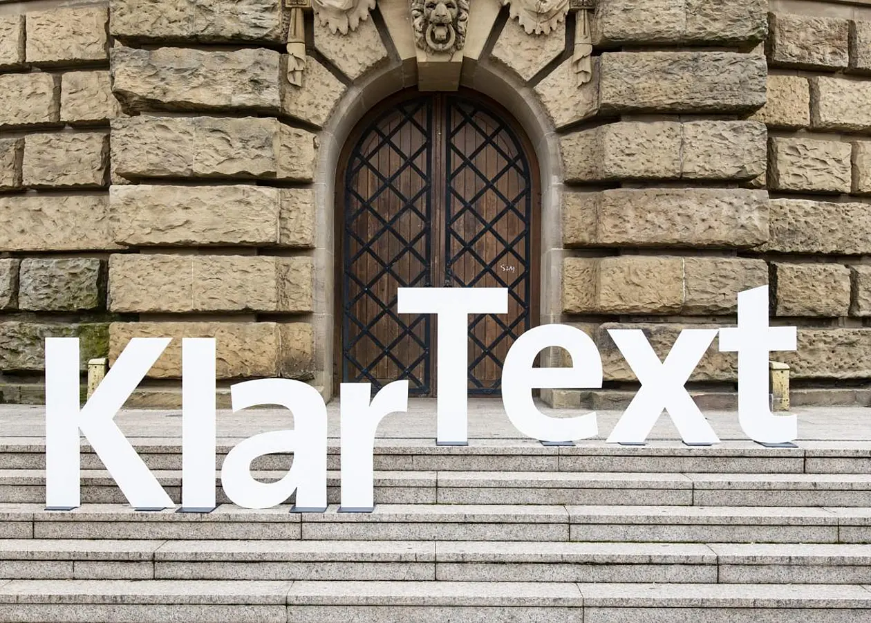 Buchstabenfolge "KlarText" aufgestellt auf Treppenstufen vor Holztür in Gebäude