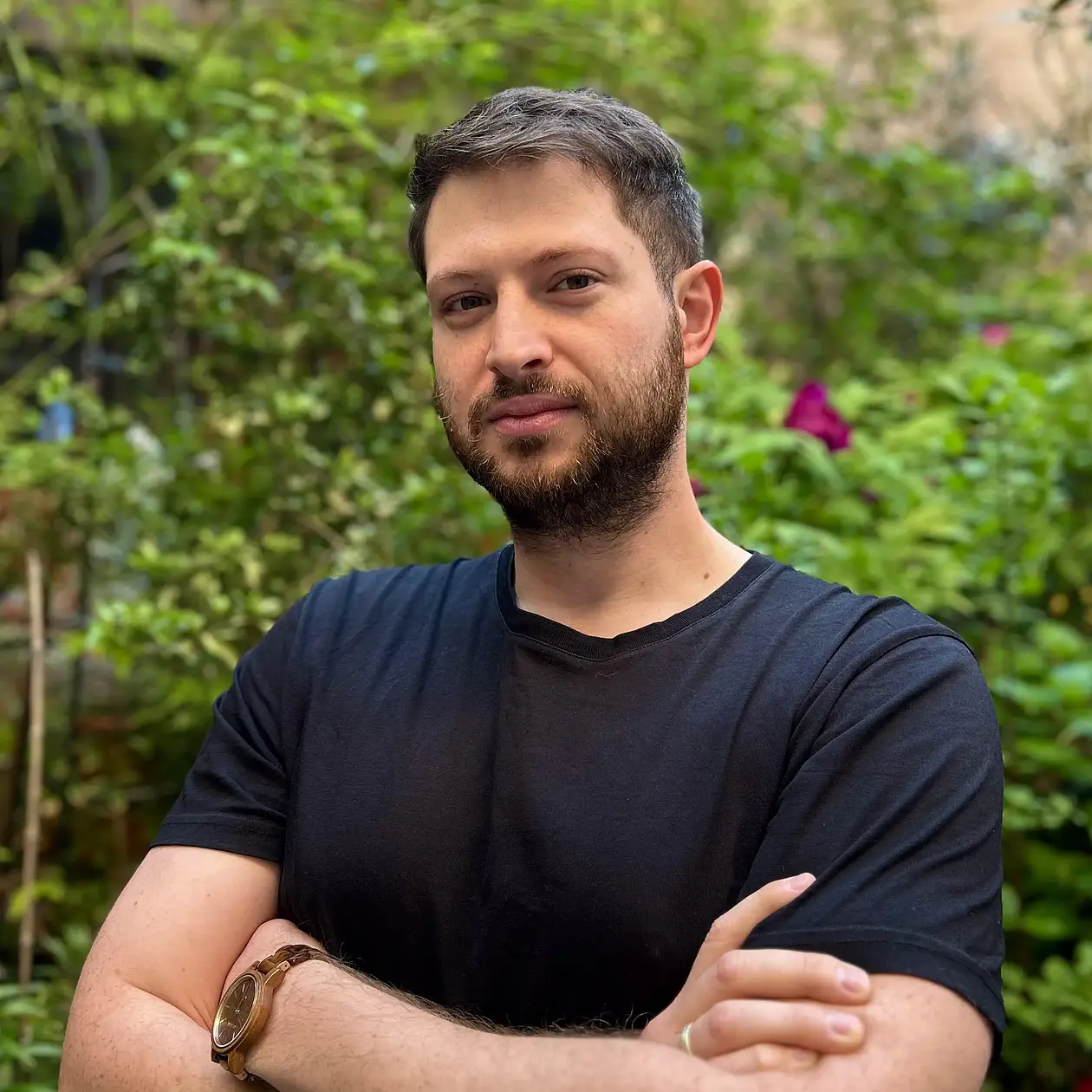 A photo of Dennis Bath, a white man standing in front of greenery wearing a dark blue short sleeve short. He faces the camera with crossed arms, and has dark brown short hair and a short beard. 