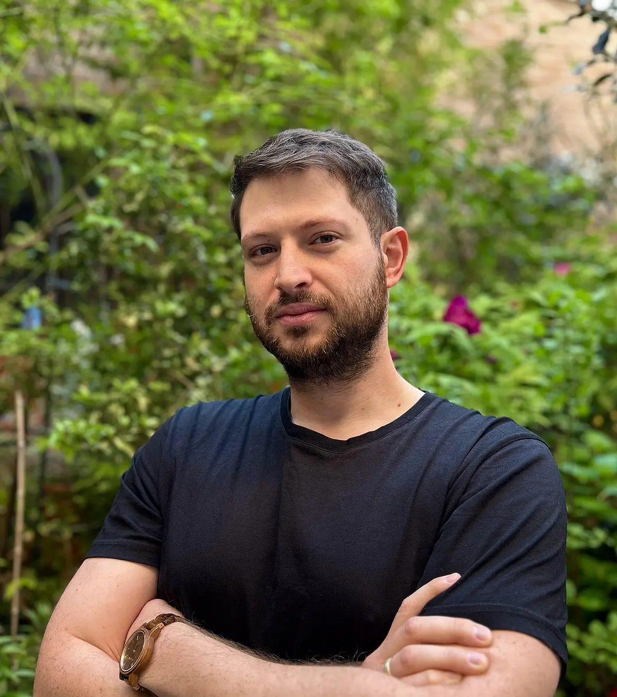 A photo of Dennis Batz, a white man standing in front of greenery wearing a dark blue short sleeve short. He faces the camera with crossed arms, and has dark brown short hair and a short beard. 