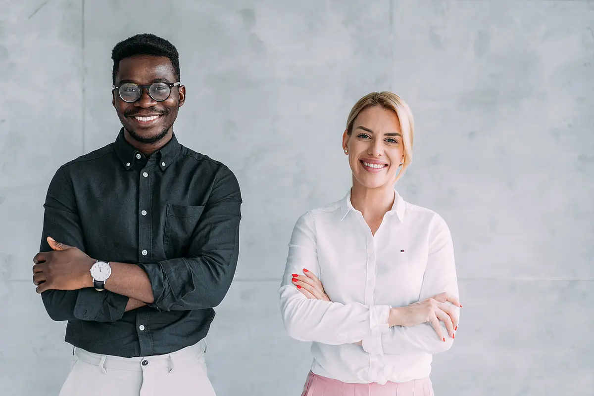Photo of a young black man and a young white woman dressed casual business, standing next to each other and smiling into the camera