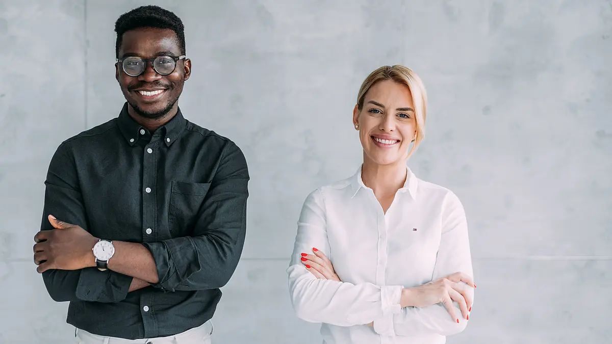 Foto zeigt einen schwarzen jungen Mann und eine weiße junge Frau im Casual Business Outfit, die neben einander stehen und in die Kamera lächeln