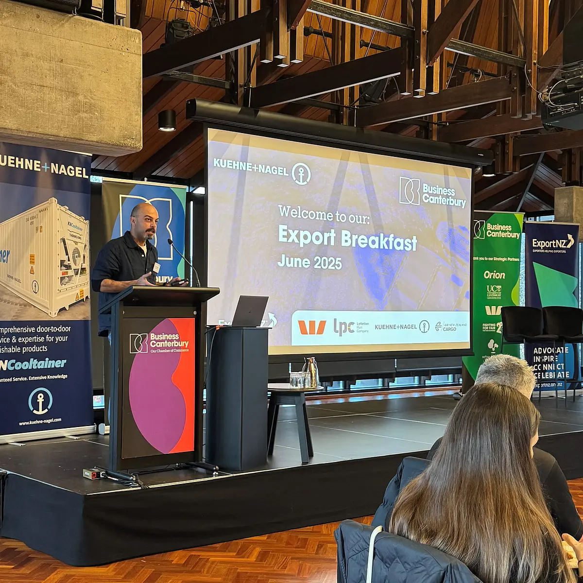 Dr. Onur Koska speaking on stage at the Export Breakfast event. A large screen behind him displays a welcome message for attendees.
