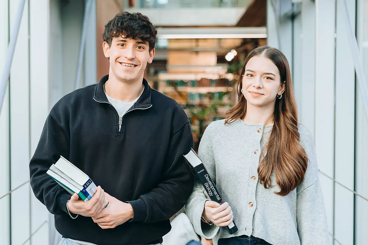 A male student and a female student are walking down a corridor, carrying books and smiling.