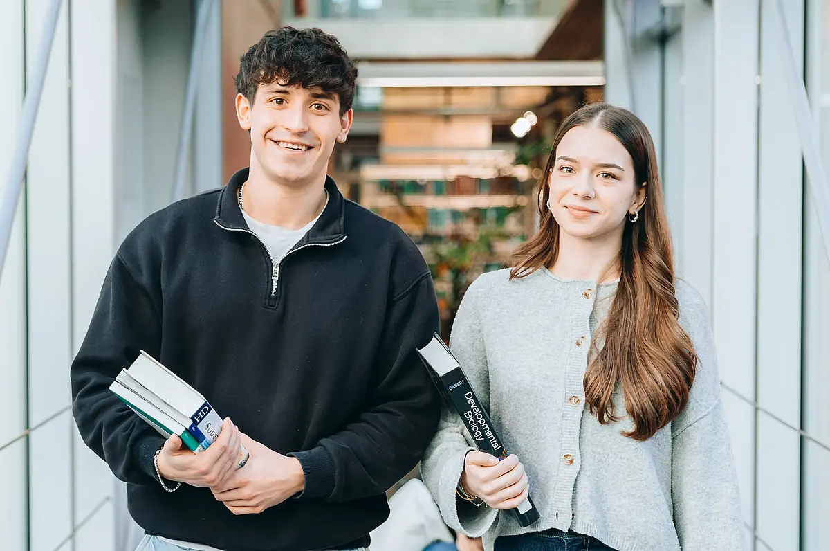 Ein Student und eine Studentin laufen einen Gang entlang, haben Bücher in der Hand und lächeln.