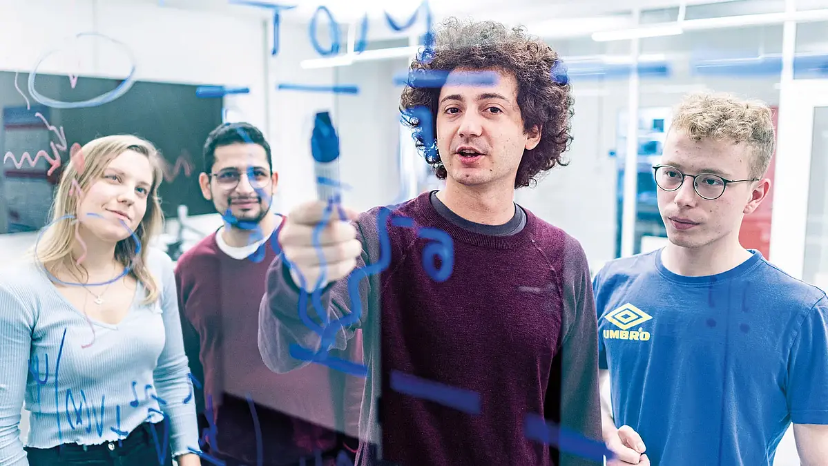 Vier junge Forschende stehen an einer Glaswand und diskutieren die physikalischen Formeln darauf. Physik Schulpatenschaften