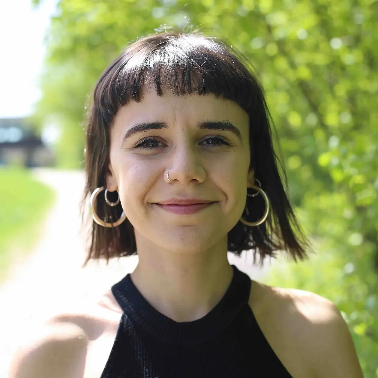 A photo of Cecilia Padilla-Iglesias, a white woman with dark brown hair ending at chin length and with a fringe, wearing a black strap top, facing the camera with a confident smile, standing outside in a green surrounding.