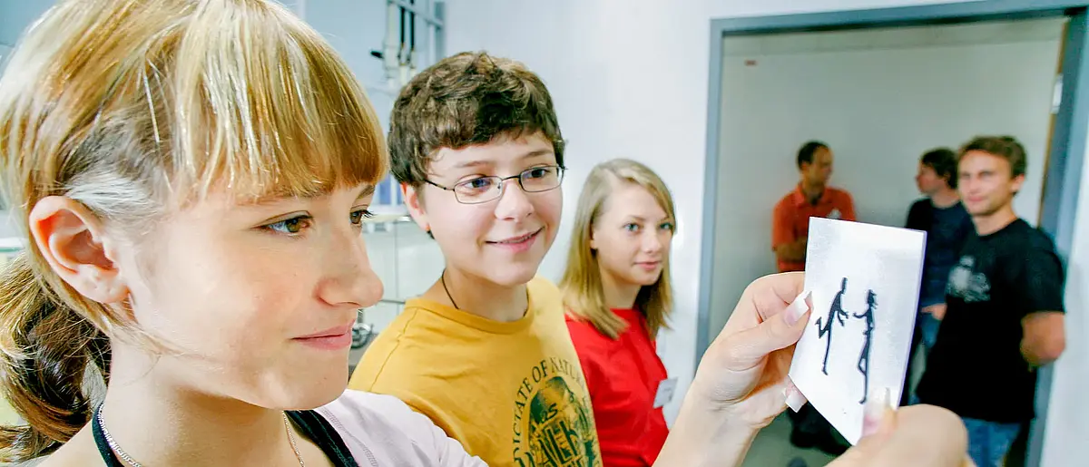 Mehrere Schüler stehen in einem Seminarraum, das Mädchen im Vordergrund blickt auf eine schwarz-weiße Zeichnung.