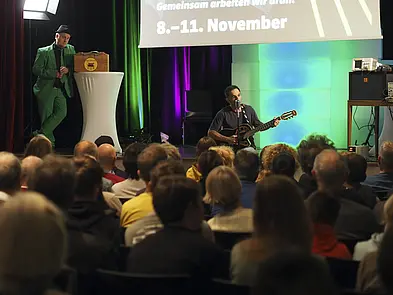 Blick auf Bühne über Publikum, auf der Bühne Mann im grünen Anzug und Mann mit Gitarre
