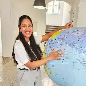 A young woman stands in the hallways of a university building. She smiles into the camera and holds a large inflatable globe.