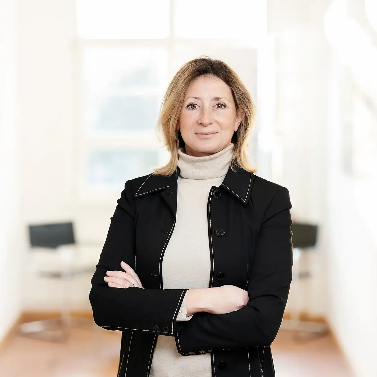 Photo of Professor Doctor Katerina Harvati, standing in a hallway with a light brown floor and white walls. She is a white woman with light brown hair down to her shoulders, wearing a black blazer and an off white blouse, facing the camera with crossed arms. 