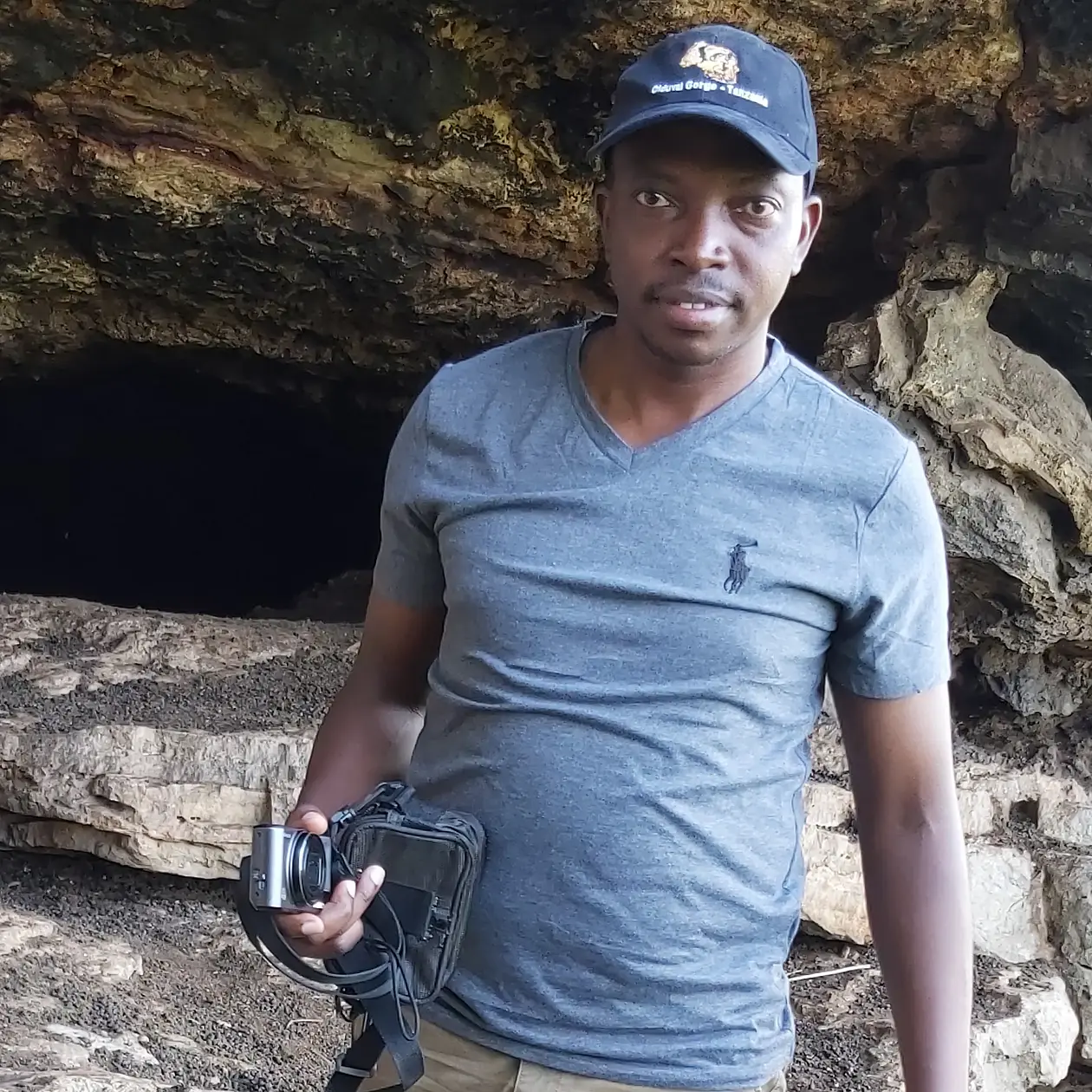 A photo of Edward Masongo, a black man wearing a basecamp and a light blue shirt in front of a cave entrance, facing the camera. 
