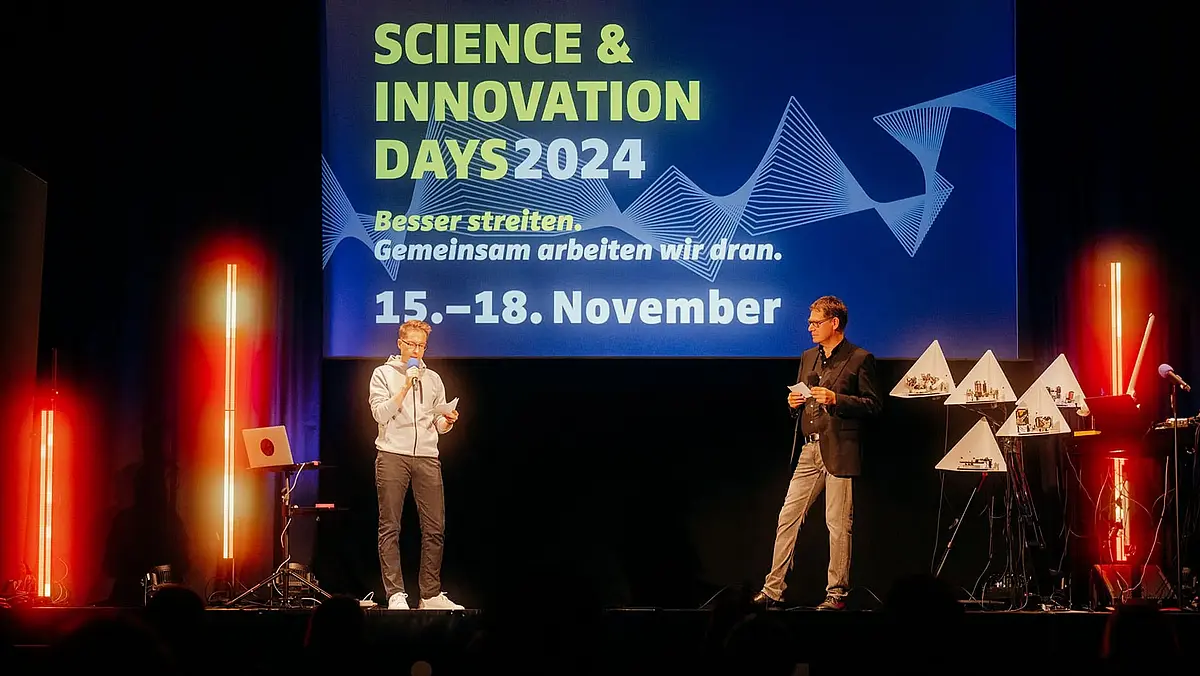Zwei Männer stehen vor einem Bildschirm mit dem Schriftzug Science & Innovation Days auf ein bunt beleuchteten Bühne und begrüßen das Publikum.