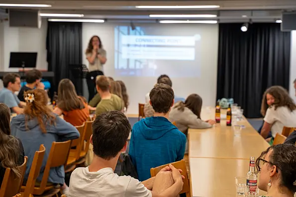 Multiple students sitting at tables listening to a woman who is standing on a stage and speaking into a microphone
