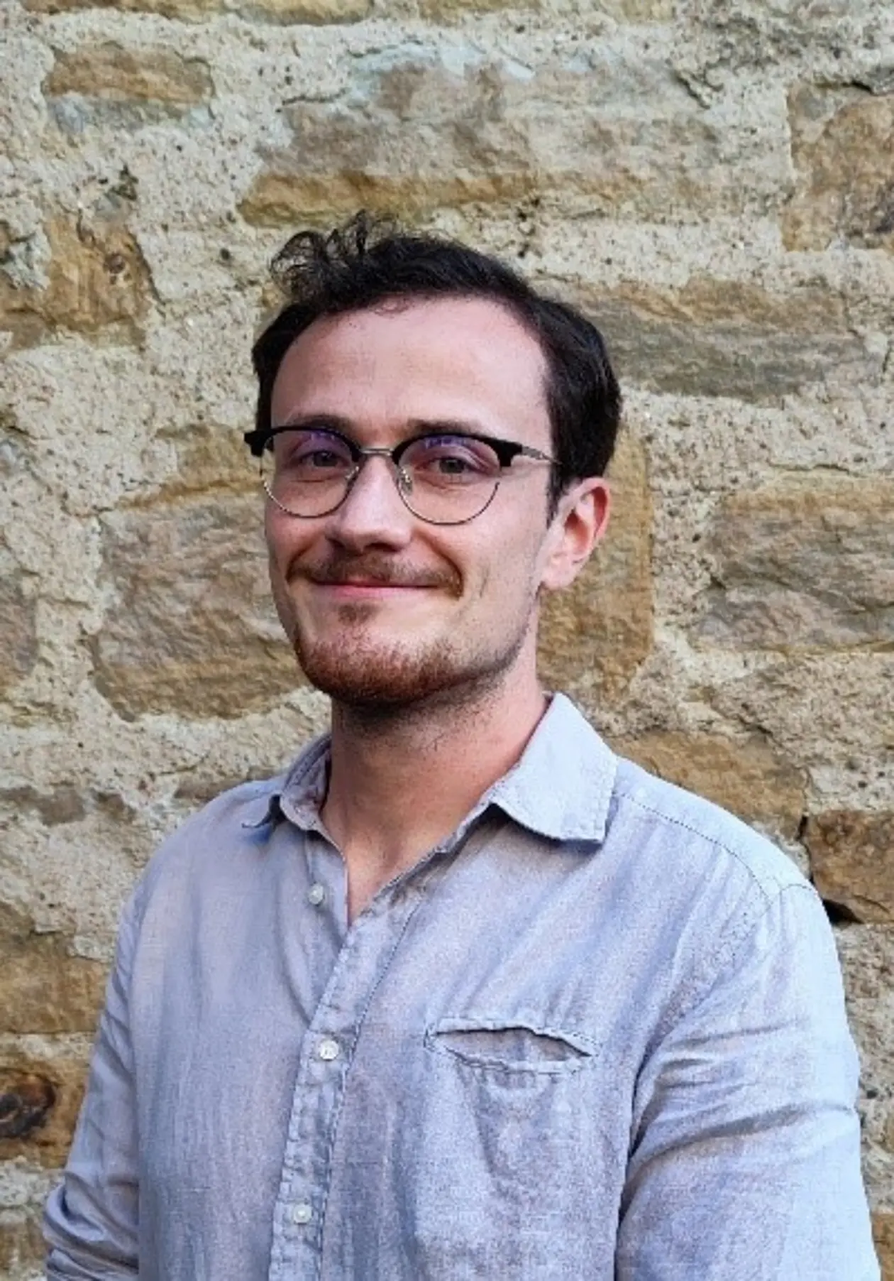Portraitfoto von Moritz Foth, weißer Mann mit dunklen Haaren, Brille und Bart. Er lächelt und trägt ein hellblaues Hemd.