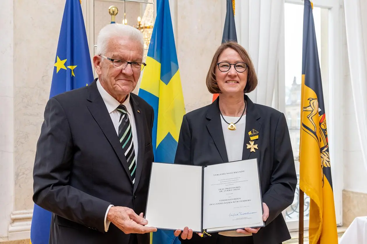 Foto von Kretschmann und Cress vor Fahnen, beide halten und zeigen eine doppelseitige Urkunde 