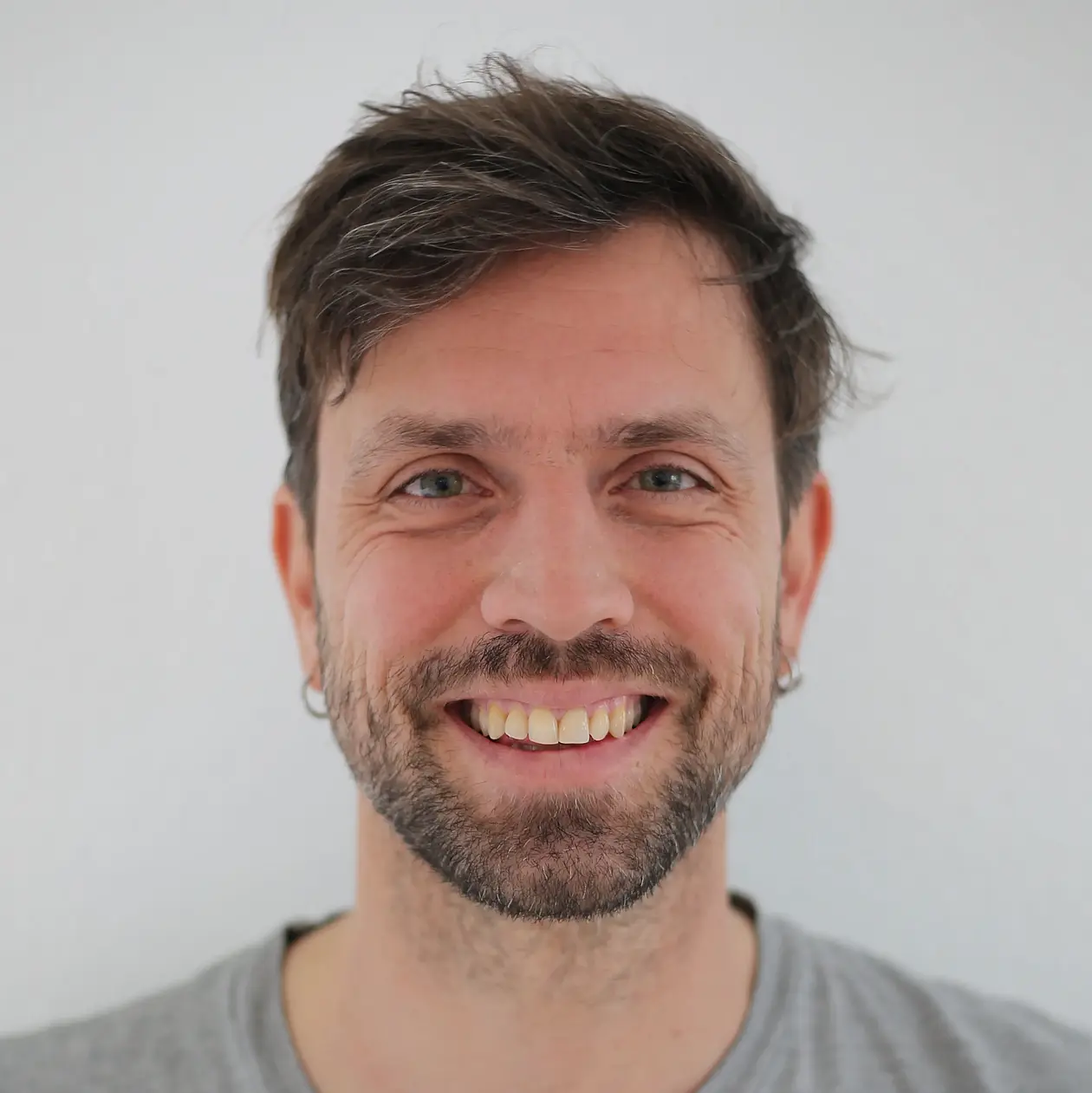 A portrait photo of Benjamin LAubi, a white man with shaggy dark brown hair and beard stubbles, smiling at the camera, wearing a grey T-Shirt. 