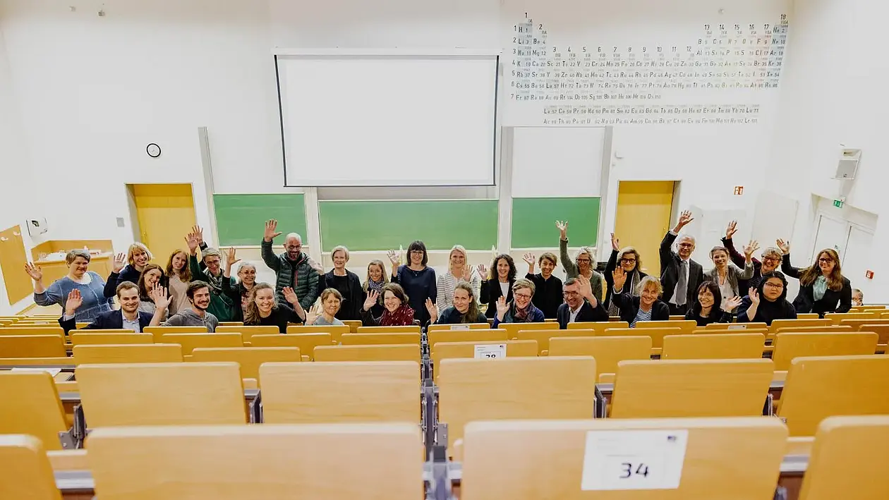 Gruppenbild im Hörsaal, Blick von hinteren leeren Stuhlreihen nach unten, die Gruppe winkt in die Kamera