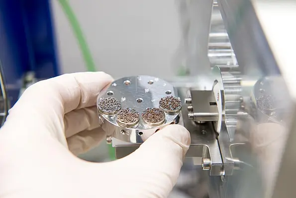 The picture shows the hand of a scientist inserting sand grain samples on a round carrier into a scanning electron microscope. 