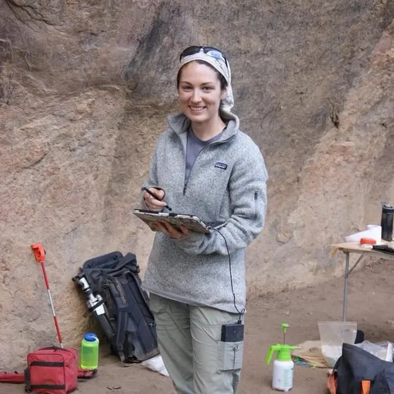 Dr. Elizabeth Grace Veatch is standing in a fieldwork site, turning towards the camera and smiling. She is a white woman wearing a light grey headscarf over dark hair, and a light grey fleece sweater. There is a sediment profile in the background and several bags and container. 