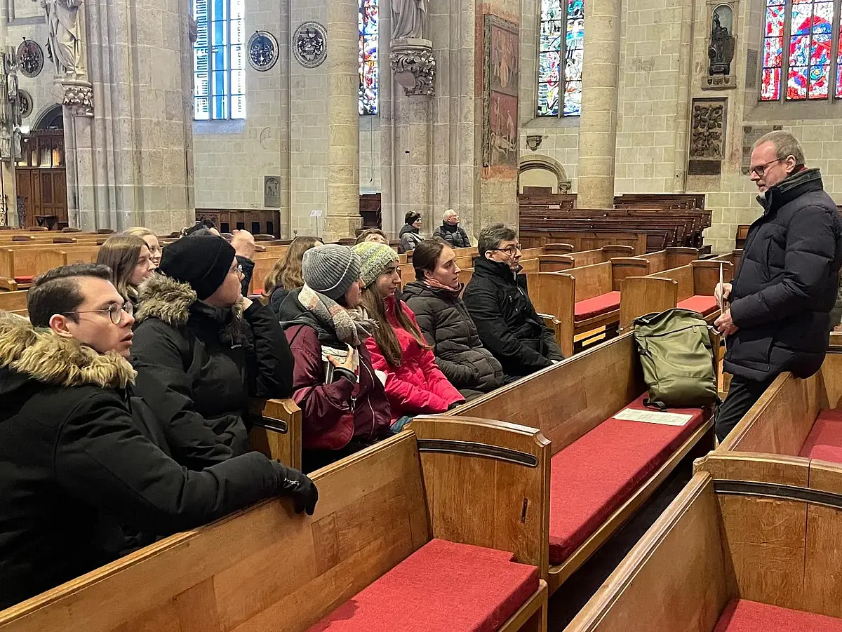 Auf dem Bild ist ein knappes Duzend Studierende zu sehen, die im Ulmer Münster (einer gotischen Kirche) sitzen. Ihnen gegenüber steht Prof. Dr. Andreas Holzem und spricht zu den Studierenden. Alle Personen sind in die dicke Jacken und Mäntel gekleidet und tragen teilweise Mützen.