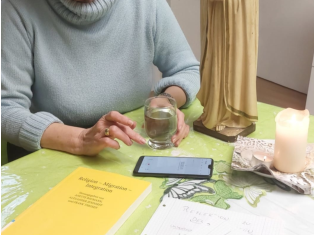 Eine Frau sitzt neben einem Tisch, auf das ein Smartphone liegt, sowie eine Kerze und ein Buch mit dem Titel "Religion - Migration - Integration". Sie tippt etwas auf dem Handy mit der rechten Hand und hält ein Glas Wasser mit der linken.