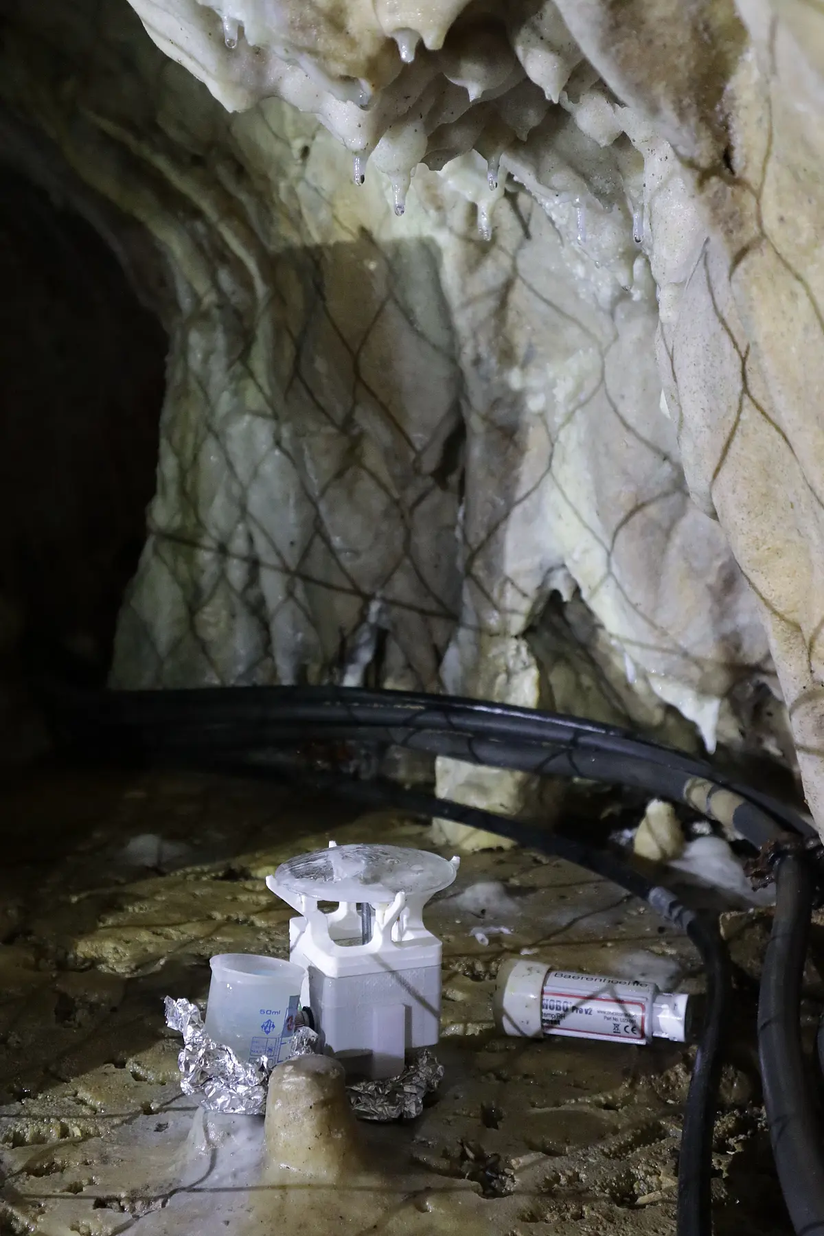 Ein Tropfwasser- und Temperaturlogger in einer Höhle neben einem Becher. Rundherum verlaufen schwarze Schläuche. Am oberen Bildrand sind Tropfsteine, an denen sich Wassertropfen bilden, zu sehen.