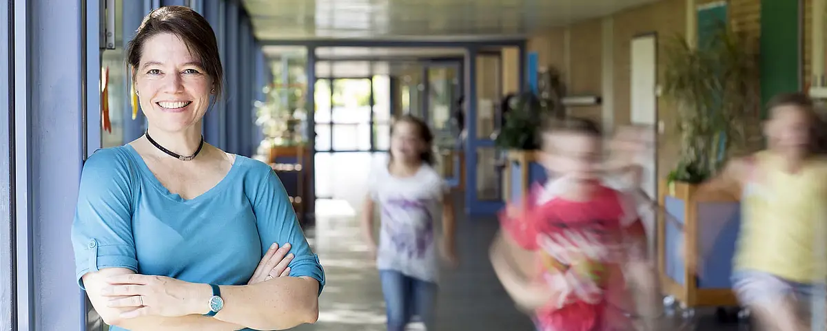 Eine Lehrerin steht in einem Schulflur, sie lächelt in die Kamera, ihre Arme sind überkreuzt. Im Hintergrund rennen Kinder.