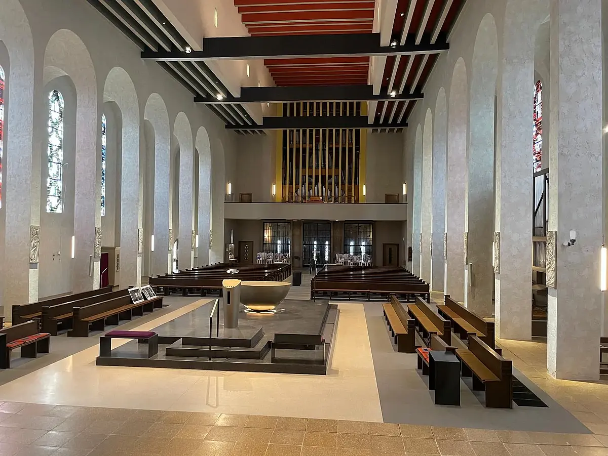 Zu sehen ist ein Kircheninnenraum. Der Blick richtet sich von hinter weit hinter dem Altarraum gen Eingang und Orgel. Besonders die sehr dunklen Holzbänke stechen im Bild hervor. Der Altarraum ist etwas erhoben und steht auf grauem Stein. Zwischen Fensterfront und Hauptschiff erstrecken sich kleinere, längliche Seitenschiffe, die durch hellgraue Kaskaden abgesetzt sind. An jeder Säule befindet sich eine Kreuzwegstation aus Eisen.