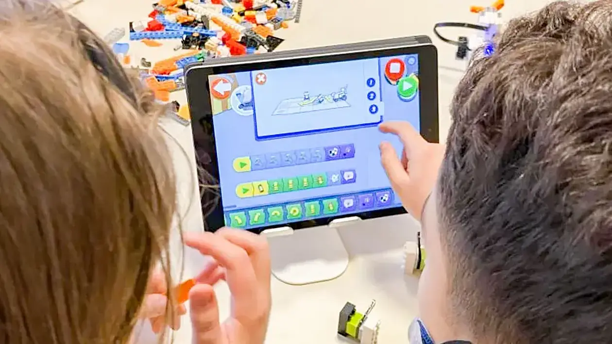 Several children are sitting at a table, working with LEGO bricks and a tablet. There are numerous building pieces on the table, as well as a partially assembled model. A graphical programming interface for a LEGO robot is displayed on the tablet.