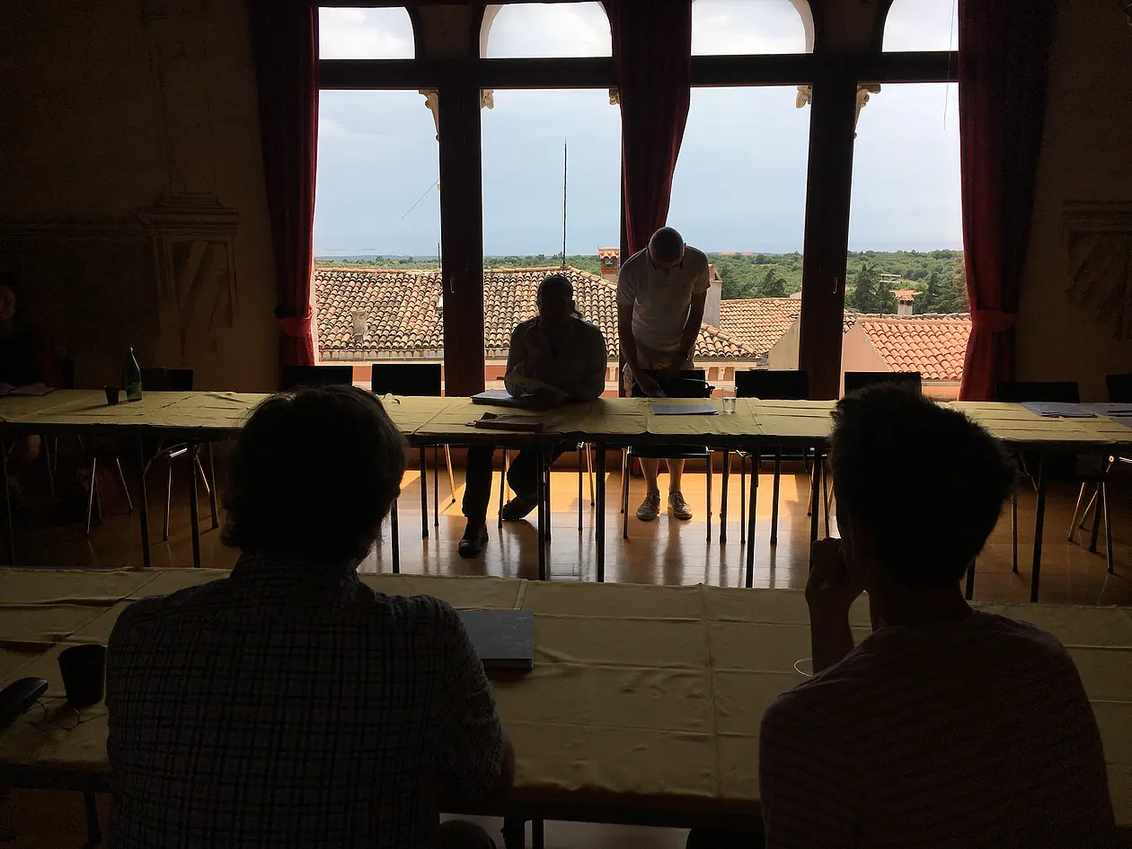 Ausblick aus einem vier mit Sprossen unterteilten hohen Fenstern, davor sitzt eine Person, ebenso im Vordergrund. Das Bild ist von einer Konferenz in Bale aufgenommen worden. 