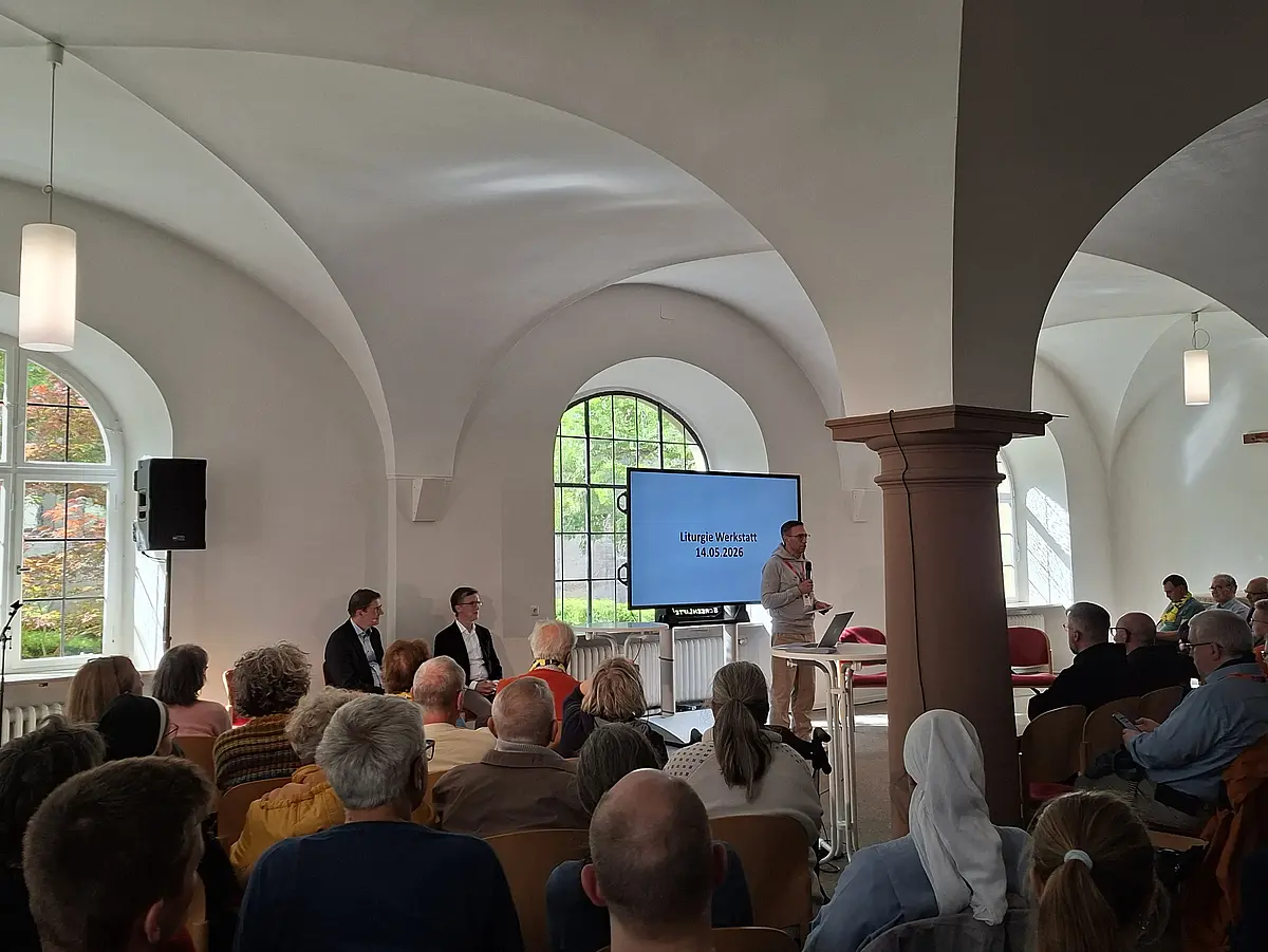 Das Bild ist in einem hellen Saal aufgenommen worden. Mittig rechts steht eine massive Säule. Der Hintergrund ist von großen Fenstern eingenommen durch die viel Licht dringt. Am unteren Bildrand ist das Publikum zu sehen, das sich in Richtung der Fenster bzw. des Flatscreens ausgerichtet hat. Vor dem Flatscreen steht Prof. Martin Stuflesser. Auf dem Bildschirm steht "Liturgie Werkstatt 14.05.2026".